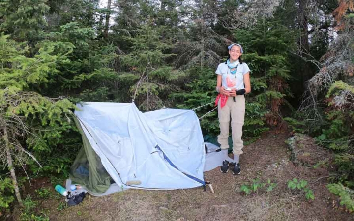 an outward bound student stands beside their tarp shelter in a wooded area 